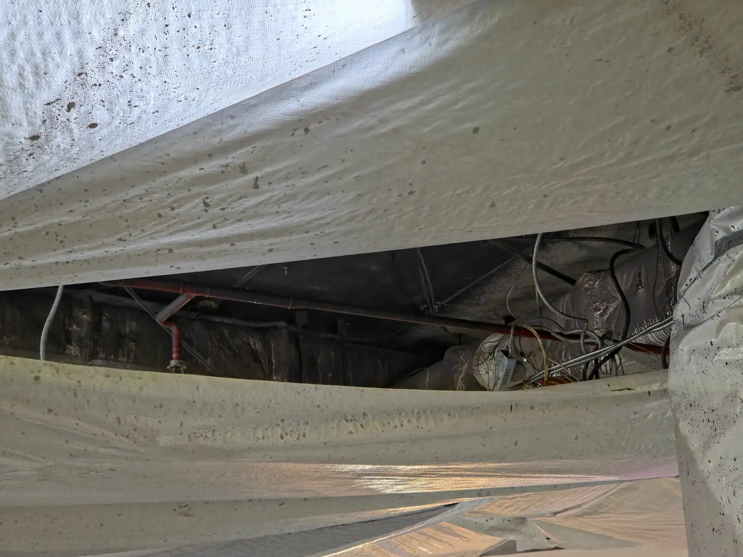View inside a building crawl space showing a white vapor barrier covering the ground and utility pipes/wires overhead